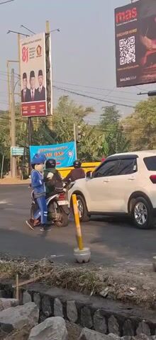 A person in a blue mascot costume, resembling a cartoon character, stands next to a white SUV. The mascot is holding something in its hands and appears to be interacting with the driver of the SUV. Two people are on a motorcycle behind the mascot. In the background, there are billboards, traffic lights, and trees. The scene appears to be an urban street during the daytime.