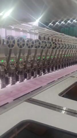 A close-up shot shows multiple industrial embroidery machines in operation. The machines are white with several green indicator lights visible. Each machine has spools of thread above it, and below the embroidery heads, the machines are stitching a delicate pink fabric with a lace-like pattern. The video pans slowly across the row of machines, revealing more spools and more sections of the embroidered fabric. The overall impression is one of automated textile production.