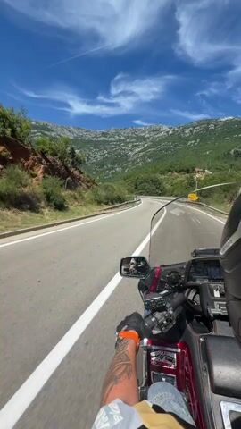 The video is shot from the perspective of someone riding a motorcycle. The rider's hands, wearing an orange glove and a black glove, are visible gripping the handlebars. A tattoo is visible on the rider's left arm. The motorcycle appears to be a large touring model, possibly a Honda Gold Wing, with a windshield. The road is asphalt and curves to the right, with white lane markings. A metal guardrail lines the left side of the road. In the distance, a mountainous landscape is visible, with green vegetation and rocky slopes. The sky is blue with some wispy white clouds. A yellow sign is visible on the right side of the road in the distance. The overall impression is a scenic motorcycle ride on a sunny day.