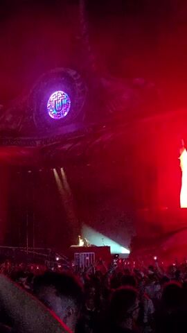 This is a first-person perspective video of a concert. The viewpoint is from within a crowd of people, looking towards a stage. The stage has a large, ornate structure above it that resembles a dragon's head. There are red lights illuminating the scene, and some smoke or fog is present. In the center of the frame, suspended in the air, is a circular object with a geometric, colorful pattern. People in the crowd have their hands raised. The overall atmosphere is one of excitement and energy, typical of a live music event.