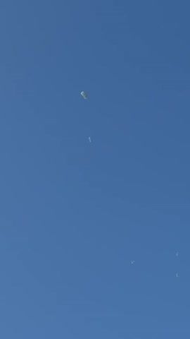 The video shows a clear blue sky with several parachutes falling. The parachutes are small and white, and they appear to be descending at a steady pace. There are no people visible in the video, nor are there any buildings or other structures. The environment appears to be open and unpopulated.
