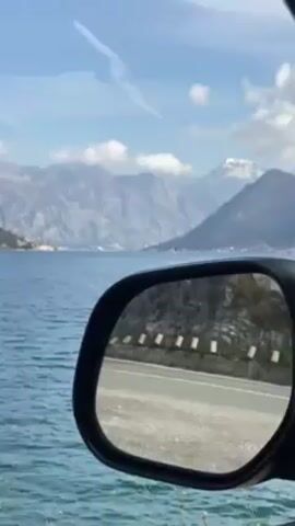 The video starts with a view out the side mirror of a car showing a blue body of water with distant mountains on the opposite shore. A motorcycle passes in the side mirror. The view pans out and shows a larger view of the water and the mountains on the opposite shore. The water has small ripples. The mountains are gray and rocky with some greenery. The sky is blue with some white clouds.