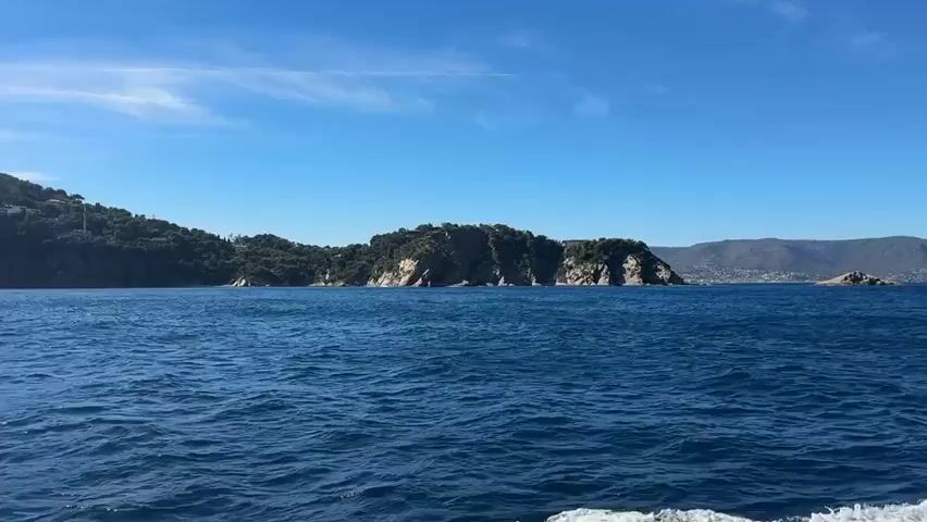 This is a video taken from a boat on a sunny day. The boat is moving through blue water, creating a wake at the bottom right of the frame. In the distance, there is a rugged, tree-covered island with cliffs and rocky shores. Beyond the island, there are hills with some buildings visible in the distance, suggesting a coastal town or city. The sky is a clear, bright blue with a few wispy white clouds. The overall impression is of a scenic coastal view from the sea.