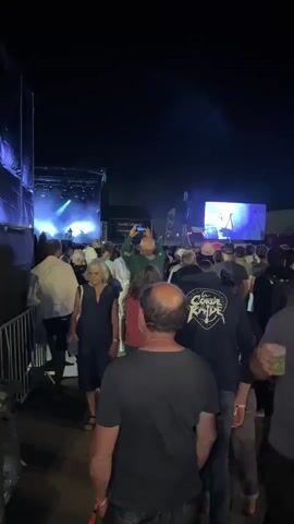 A low-angle, slightly shaky, vertical video shows a crowd of people gathered at an outdoor concert at night. The stage is visible in the background with bright lights and a large screen displaying performers. People are walking and standing, some holding drinks and phones. The atmosphere appears lively and energetic. The camera is moving, capturing candid moments of concert-goers. The lighting is dim, with stage lights and screens illuminating the scene. The overall impression is of a vibrant concert event with a diverse audience enjoying the music and ambiance.