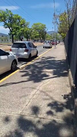 The video shows a sidewalk on a sunny day with a gray concrete surface. The sidewalk is bordered by a gray fence on the right and a street on the left. Cars are parked along the street. The video is still and there are no people visible. Tree shadows cover the sidewalk. The sky is blue with some light clouds. There are green trees in the background and a mountain range in the far distance.