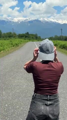 A lone female figure is captured from behind, walking away from the camera down the center of an asphalt road. She is wearing a grey baseball cap, which she adjusts briefly at the beginning of the clip, a fitted dark reddish-brown short-sleeved top, dark grey/black high-waisted denim shorts with a frayed hem, and white sneakers with black accents.

The road, marked with a single white dashed line down its middle, stretches straight ahead and gently curves, leading towards a majestic range of distant, snow-capped mountains. Lush green vegetation, possibly ferns or dense bushes, lines both sides of the road. Overhead, the sky is a vibrant blue with scattered white cumulus clouds, indicating a bright, sunny day. The scene appears to be a rural or remote area, potentially within Akwaskia tshyts, Abkhazia, offering a breathtaking natural landscape. Power lines and utility poles are faintly visible on the left side of the road in the distance. The overall atmosphere is one of peaceful exploration and appreciation for the scenic surroundings.