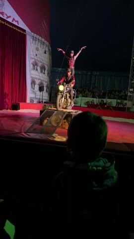A child's head is visible in the foreground of a video, turned towards a circus performance. A man in a red and black costume is seated on a motorcycle in the center of the stage, with a woman standing on his shoulders. She is also dressed in red and black, and her arms are raised. The motorcycle is on a platform with ramps. The background shows a painted backdrop resembling a building with arches, and red curtains on the left. An audience can be seen in the dimly lit background under the blue tent.