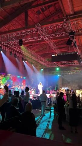 A concert is being held in a large indoor venue. On stage, a band is performing, with spotlights illuminating the performers and the audience. The audience is spread out, with some people standing and watching the performance, while others are seated at tables. The venue itself appears to be a converted industrial building, with exposed beams and brick walls. The lighting is dynamic, with colored spotlights and beams of light creating a festive atmosphere. There is a dance floor in front of the stage where some audience members are dancing.