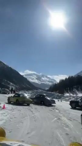 The video shows a group of vintage cars parked on a snowy surface, possibly a frozen lake or a road covered in snow. The sun is shining brightly, and the sky is clear blue. The landscape includes snow-covered mountains and trees, suggesting a winter or alpine setting. The cars are a mix of classic models, including several Porsches, and they appear to be part of a rally or gathering. The camera moves slightly, showing the cars from different angles and highlighting the snowy environment. The overall scene depicts a picturesque winter setting with classic cars against a backdrop of snow-covered mountains.