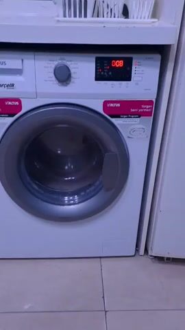The video shows a close-up view of a white Altus brand washing machine with a silver door. The machine is standing on a tiled floor, and there is a white surface above it, possibly a shelf or countertop, with some items on it that are out of focus. The washing machine has a digital display that shows "0:08" and several small indicator lights. There are also control knobs and buttons on the front panel. Two pink labels are attached to the front of the machine, one on the left and one on the right, with text in Turkish. The overall environment appears to be a laundry room or a similar domestic setting. The video is static, with no movement or activity captured.