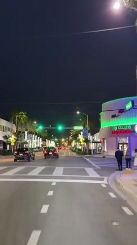 The video shows a nighttime view of a street at an intersection. Cars are driving on the road, and streetlights are illuminated. Palm trees line the sidewalks, and buildings with bright, colorful lights are visible on both sides of the street. The sky is dark, indicating it is nighttime. There are crosswalks and traffic signals at the intersection. One person is walking across the street. The overall scene depicts a lively, urban environment at night.