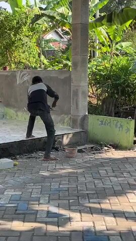 A person is standing on a raised concrete platform, facing away from the camera. They are wearing a dark long-sleeved shirt with lighter colored sleeves and dark pants. Their left arm is bent and extended forward. The person appears to be working on a wall that is partially tiled. The background consists of lush green vegetation, including banana trees. The foreground shows a paved surface with scattered construction debris, including bricks, a bucket, and rolls of wire.