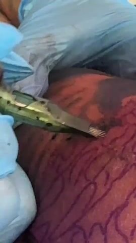 A close-up video shows a tattoo artist's gloved hands holding a tattoo gun. The tattoo gun is being used to apply ink to a person's skin, which is a reddish-brown color and has faint purple lines drawn on it, presumably as a stencil. The tattoo gun appears to have multiple needles. The background is out of focus, but appears to be blue cloth.