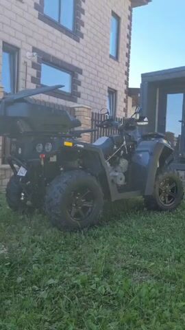 A dark gray ATV with a black cargo box is parked on a patch of green grass in front of a light brown brick house. The ATV has four large black tires with aggressive tread patterns. The rear of the ATV has multiple lights and what appears to be a license plate. A small fence is behind the ATV, and a portion of the house's foundation is visible. The sky is clear and blue. The camera is focused on the ATV and its surroundings.