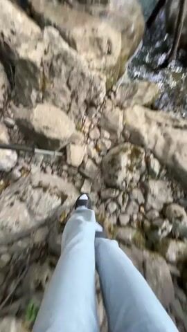 The video shows a person walking on rocks towards a small waterfall in a wooded area. The person is wearing light blue jeans and black shoes. The waterfall cascades over a rocky cliff into a small pool. Trees and foliage surround the area. A person wearing a cap is visible briefly near the end of the video, reaching towards the water.