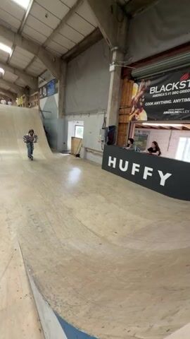 A young person wearing a helmet and striped top is riding a scooter in an indoor skate park. The park features wooden ramps and walls. The person performs a jump, catching air, and lands back on the ramp. The park is in a large indoor space with a high ceiling and visible support beams. A banner with "HUFFY" is displayed on a wall. Other signs are visible in the background. There are people watching from behind the banner. The camera follows the action, maintaining a close to medium shot of the scooter rider. The lighting is bright.