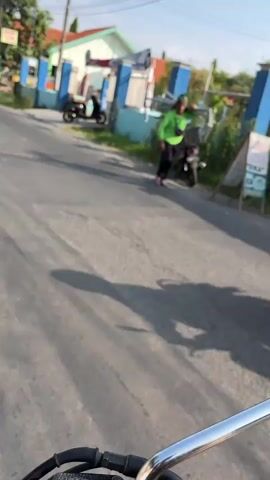 The media file shows a street scene in Sidoarjo, East Java. The foreground is dominated by the handlebars and front of a motorcycle, indicating the perspective of someone riding or filming from a motorcycle. In the middle ground, a person wearing a bright green shirt and dark pants is standing next to a parked motorcycle. They are leaning on the handlebars and appear to be looking off-camera. Behind this person, a light blue fence and building structures are visible, suggesting a residential or commercial area. Another motorcycle is parked further down the street.

The time of day appears to be late afternoon or early evening, as indicated by the long shadows cast on the asphalt road, creating a dappled effect. The sunlight suggests a clear or partly cloudy sky. The overall mood seems relaxed and everyday, with no overt signs of distress or excitement from the person in the green shirt. The environment appears to be an urban or semi-urban setting, with paved roads and buildings characteristic of a Southeast Asian locale. There are no discernible emotions conveyed by the person, and they are the only clearly identifiable person in the frame. The visible text on a sign to the right of the frame is too blurry to be read.
