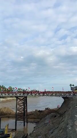 The camera pans around showing a bridge that is decorated with many red and white flags. The bridge spans across a river with muddy brown water. There are construction vehicles below the bridge and construction work being done on the river bank. The sky above is cloudy. There are cars and people on the bridge.