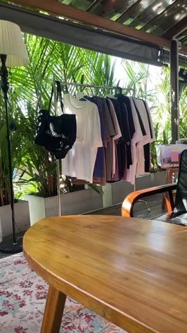 A clothing rack with various colored shirts is displayed outdoors. A black tote bag hangs on the left side of the rack, and a tall floor lamp is positioned to the left of the rack. In the foreground, a wooden table and a rug are visible, with lush green foliage forming the backdrop. The scene appears to be a cafe or an outdoor retail space.