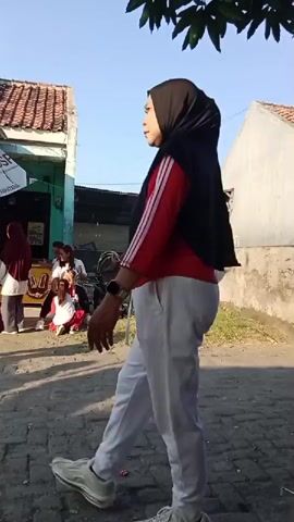 In the first segment, a child in a red t-shirt and a woman in a hijab and red-striped jacket walk from right to left. The child is drinking from a plastic cup. In the second segment, a woman wearing a white t-shirt with black stripes and the number 88, along with black pants, walks from right to left, holding a blue umbrella. In the third segment, a young child in a red and white shirt and light-colored pants walks from right to left. In the background, two children sit with a stroller. A flag with red and white stripes is visible. The environment appears to be an outdoor setting with houses and a paved walkway. In the fourth segment, a young person in a black baseball cap, a red and black checkered t-shirt, and black shorts walks from right to left, gesturing with their hands. In the fifth segment, a child in a white t-shirt stands facing the camera, holding a red and white object in their hands. A person in a red and black checkered t-shirt is visible in the background.