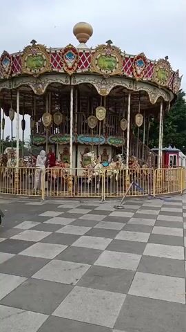 A carousel ride is shown from the outside, with people standing around it. The carousel is decorated with gold accents, and has horses for people to ride on. The ground in front of the carousel is a checkerboard pattern of light and dark gray tiles. There is a fence around the carousel.