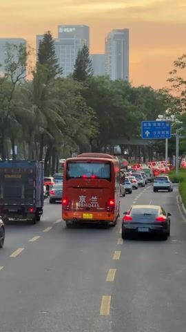 The video shows a scene of traffic on a road in what appears to be a city in China, at dusk. The camera is positioned behind a line of cars, facing forward. There are various vehicles on the road, including cars, a truck, and a red bus with Chinese writing on the back. The sky is a pale orange, indicating either sunrise or sunset. Trees line the side of the road, and in the distance, there are several tall buildings. A road sign is visible, with Chinese characters and arrows. The traffic appears to be slow-moving or at a standstill.