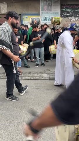 The media file shows a street scene in Babol, Iran, likely during a religious procession or commemoration. The foreground features a person's arm and shoulder, blurred due to the close-up perspective and focus on the background action.

In the middle ground, a group of people are gathered on a street, with buildings lining the background. Several individuals are actively playing drums, which are large and cylindrical, some with yellow drumheads. One man on the left wears a black t-shirt and jeans, and has a patterned scarf draped around his neck, holding a long, curved horn-like object. To his right, another man in a white robe and a patterned headscarf is playing a large drum. Other people in the crowd are also holding drums, suggesting a musical element to the event. The participants are mostly men, but there are women and children present as well, some wearing headscarves. The atmosphere appears to be one of solemnity or shared cultural activity.

In the background, the buildings have signs with Arabic script, indicating the location. One prominent poster on a building shows a portrait of a man, possibly a religious figure. The sky is not visible, but the lighting suggests it is daytime, possibly late morning or afternoon. The scene takes place on a paved street with a curb. The overall impression is of a community engaged in a traditional observance.