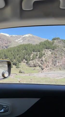 The video, taken from inside a car, showcases a mountainous landscape. The window frames the view, with the car's interior visible at the top and side. The landscape includes a hillside densely populated with green trees. The hill appears rocky with exposed areas of dirt and stone. In the background, a larger mountain range is visible, partially covered in snow. The sky is clear and blue. Power lines are present on the top of the hill. The car is moving, as the scenery shifts.