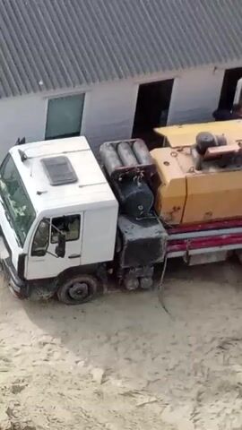The video shows a construction site. A white truck with a yellow concrete pump attached is parked near a small building with the logo "LVIV BUD" on it. A person is seen inside the building. A concrete mixer truck is positioned to pour concrete into the pump. The concrete is then pumped through a hose to an elevated platform where several construction workers are spreading the concrete. A walkway is visible, leading from the building to the elevated platform. A blue portable toilet is also present on the site.