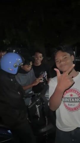 A group of four young men and one woman are gathered around a motorcycle at night. The woman is wearing a blue helmet and a dark hooded jacket, and she is smiling and waving. One of the men, wearing a white t-shirt with a graphic on it, is pointing at the camera and smiling. Another man, wearing a dark t-shirt, is holding a phone and appears to be showing something to the others. The scene is dimly lit, with trees and some indistinct background lights visible. The overall atmosphere appears to be casual and friendly.