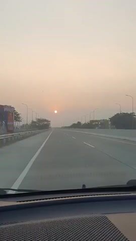 A first-person perspective video shows a car driving on a highway at sunset. The sun is visible in the distance, low on the horizon, casting a warm glow over the sky. The highway is mostly empty, with a few streetlights lining the sides. Trees and some structures are visible in the background. The interior of the car, including the dashboard, is partially visible in the foreground. The video appears to be a time-lapse, as the sun's position and the surrounding scenery change rapidly.