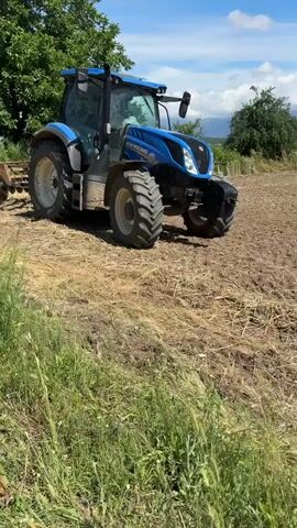 A blue New Holland tractor is parked in a field on a sunny day. The tractor is facing the camera and is positioned on dry, matted grass and dirt. The front of the tractor is slightly to the right of center. The tractor has large, knobby tires. A portion of a farming implement, possibly a plow or tiller, is visible attached to the rear of the tractor. In the background, there are green trees and a bright blue sky with some clouds. The field appears to be recently worked, with uneven soil. The overall scene depicts agricultural activity in an outdoor setting.
