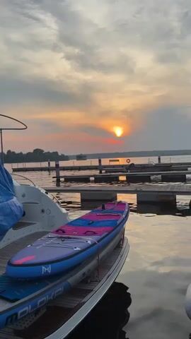 The video shows a tranquil harbor scene at sunset. The sun, a glowing orb, is partially obscured by clouds, casting a warm orange and pink hue across the sky and water. In the foreground, a paddleboard with a vibrant blue and pink design rests on the deck of a white boat. The boat is docked at a marina, with wooden piers stretching out into the calm water. Several other boats are visible in the distance, as well as a few buoys and navigational markers. The water is still, reflecting the colors of the sunset and the surrounding structures. A person is visible on the dock in the distance, but their actions are unclear. The overall atmosphere is peaceful and serene.