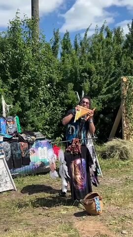 A person with long dark hair and sunglasses stands outdoors playing a pan flute. They are wearing a dark shirt and pants. In front of them is a display of t-shirts with Native American-themed designs, as well as dreamcatchers hanging from a rack. Behind the person, there is a teepee and a pile of hay. The background consists of lush green trees and a blue sky with white clouds. The ground is covered in dirt and grass. The overall scene appears to be a market or festival setting.
