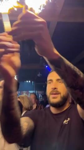A group of people celebrate in a dark bar-like setting, holding up lit sparklers. A man with tattoos on his arms holds the camera as he films. He and other people around him sing along to music. The faces of several people are visible, including a woman in glasses singing and holding a sparkler, another woman in the background, and a man with a beard singing. A disco ball reflects light near a wood and metal ceiling. Blue lights and a red light flash. Other people can be seen behind the group.