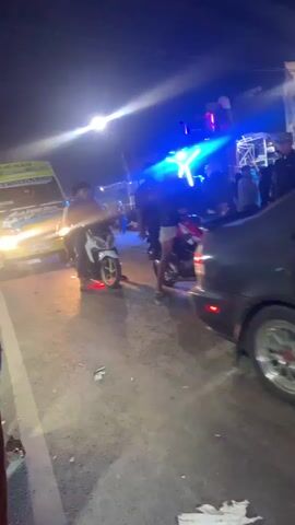 The video shows a nighttime outdoor scene with several motorcycles and a truck. There is a bright blue light illuminating a structure in the background, possibly a stage with speakers. People are visible standing around and on motorcycles. The truck is yellow and white with graphics and text on its side. One motorcycle in the foreground has red lights. The road appears to be asphalt, and there is some debris on the ground. The lighting is dim except for the bright artificial lights.