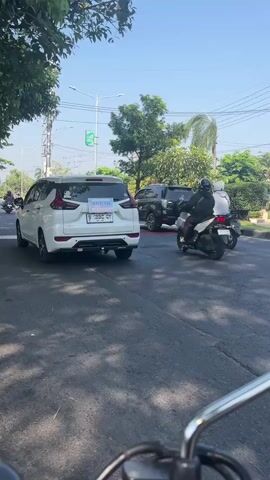 This is a first-person perspective video taken from a motorcycle in Larangan, Indonesia. The scene is a paved road with traffic. A white SUV is in the left lane, with a sign on its rear windshield. A motorcycle with two people is in front of the SUV, and another motorcycle is to the right. The rider of the video is looking forward at a red motorcycle in the right lane. The sky is clear and blue, indicating a sunny day. Trees line the side of the road, and there are utility poles and wires overhead. The time of day appears to be daytime, likely morning or afternoon. The overall mood is casual and observational. There is some text visible on the white SUV's license plate, which reads "B 1995 QY". The red motorcycle in the foreground has a license plate with the text "YN 6380 H".
