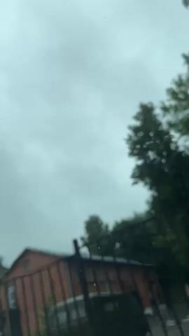The video shows a view of a brick building with a dark roof, surrounded by trees under a cloudy sky. A black metal fence is in the foreground, partially obscuring the view of the building and a vehicle parked in front of it. The camera appears to be inside a vehicle, possibly looking out through a window, as there are some water droplets visible on the surface. The overall atmosphere is overcast and appears to be daytime.