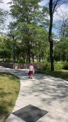 A young child wearing a red hat and a red and white outfit is riding a scooter down a paved path in a park. The child is moving away from the camera. The path is made of light-colored rectangular stones and curves to the left. To the left of the path is a grassy area. To the right of the path, there are trees with green foliage and a wooden fence. There are also some concrete benches along the path. The sky is visible in the background and appears to be cloudy. The overall environment suggests a sunny day in a park setting.