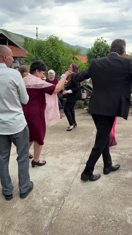 A group of people are dancing in a circle on a concrete courtyard. They hold hands, swaying and stepping to the music. Most are dressed in formal attire, including dresses and suits. A woman in a maroon dress and pink shawl is prominent in the foreground. Other figures are partially visible, including one in a blue dress, another in a pink gown with floral accents, and several men in suits. In the background, there are trees, buildings, and a stroller. The sky is overcast, suggesting an outdoor event. The scene appears to be a celebration, possibly a wedding or festive gathering.