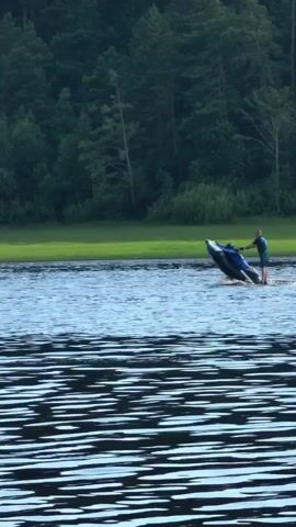 A person is riding a jet ski on a body of water. The jet ski is dark blue and black, and the rider is wearing a green t-shirt and shorts. The rider is standing on the jet ski, which is tilted up at an angle. There is a wake behind the jet ski, indicating it is moving at a good speed. The water appears to be calm, with ripples on the surface. In the background, there is a lush green grassy area and a dense forest of evergreen trees. The sky is not visible, but the lighting suggests it is daytime, possibly late afternoon or early evening, given the soft light on the trees. The scene appears peaceful and recreational. The location is identified as Sysert, Russia, which is known for its natural beauty and lakes. There is no visible text in the media file.