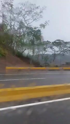 The video depicts a drive along a wet, asphalt highway on an overcast and rainy day. The camera appears to be inside a moving vehicle, looking out through a window.

**Subjects:** In the foreground, a prominent yellow concrete barrier runs alongside the road, separated by a white line. The road itself has white lane markings. Beyond the road, a yellow metal guardrail lines the edge. Various trees, some with sparse branches suggesting deciduous nature and others with full green foliage, are visible along the roadside and on distant hills.

**Activity:** The recording vehicle is moving forward. At the 0:01 mark, a yellow truck passes in the opposite direction, creating a splash from the wet road.

**Notable Details:** The weather is distinctly rainy and cloudy, with mist obscuring parts of the distant mountainous landscape. Around the 0:03 mark, a small, simple roadside building with a corrugated metal roof comes into view, accompanied by what appears to be an open-air stall or shelter next to it. A large, leafy tree stands beside these structures, and a white pickup truck is parked nearby.

**Visible Text:** On the small building, faint text "KM 3..." is visible, indicating a kilometer marker.

The scene suggests a journey through a rural or semi-rural area, possibly mountainous, under gloomy weather conditions.