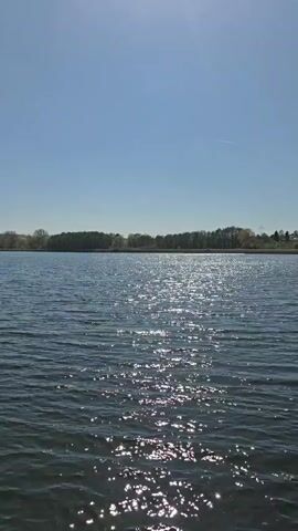The video shows a peaceful lake scene on a sunny day. The lake is calm with gentle ripples reflecting the sunlight. In the distance, trees line the horizon. The camera pans to the left, revealing a wooden dock extending into the lake. The sky is a clear blue.