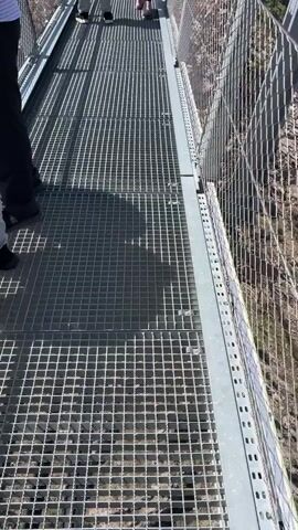 The video begins by looking down at the grated metal walkway of a suspension bridge, with people's legs visible in the foreground. The camera then pans up and moves forward along the bridge. Several people are walking ahead, including a man in a white t-shirt and khaki pants, holding a bag, and two women. The bridge is high above a rocky, mountainous landscape with sparse vegetation. The sky is clear and blue. The camera continues to move forward, offering a view of the expansive mountainous terrain and the impressive structure of the bridge.