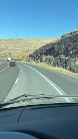 This media file captures a first-person perspective of driving on a highway under a clear, bright blue sky, suggesting a sunny daytime. The scene is arid and mountainous, with dry, golden-brown hills flanking the road. A large, dark, rocky outcrop dominates the right side of the frame, contrasting with the softer, rolling hills on the left.

The activity is vehicular travel. Several cars are visible on the road, moving in the same direction as the viewer. A white truck is further ahead, and a red car is in the lane to the left. A silver SUV with a roof rack is also in the left lane. The perspective is from inside a vehicle, looking out through the windshield, which shows windshield wipers. The dashboard of the vehicle is visible in the foreground.

A notable detail is a yellow road sign placed on the right side of the road, near the rocky outcrop. The text on the sign is legible and reads "RUNAWAY TRUCK RAMP 1 MILE". This indicates the presence of a safety feature for heavy vehicles on a downhill grade, suggesting the road may have a significant incline ahead. The location is identified as Tutuilla, United States, which aligns with the dry, hilly, and mountainous terrain depicted. The overall mood is one of travel and steady progression through a scenic, natural landscape.