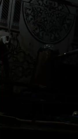 A dark video shows a copper pot on a gas stove. The pot is filled with liquid that is bubbling and about to boil over. The stove is on, indicated by the blue flames visible beneath the pot. The background appears to be a tiled wall with an ornate pattern. Some green leafy plants are visible in the foreground, suggesting a kitchen or food preparation area. The lighting is dim, casting shadows and obscuring some details.
