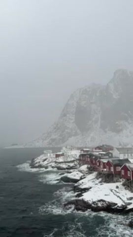 The video shows a coastal landscape in winter. Red wooden houses are built on the rocky shoreline, which is covered in snow. The sea is dark and choppy, with waves breaking against the rocks. A large, snow-covered mountain looms in the background, shrouded in mist or fog. The overall scene is grey and overcast due to the heavy snow.