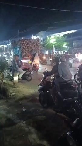 The video depicts a nighttime scene on a street, possibly in a Southeast Asian country, with a mix of people and vehicles. Several motorcycles are visible, some with riders, others parked. A truck carrying a large load of what appears to be sugarcane or bamboo is prominent in the center-left of the frame. Streetlights and various other lights illuminate the area, creating a somewhat festive or busy atmosphere. Some individuals appear to be gathered along the roadside, while others are on motorcycles, suggesting a potential event, market, or a busy thoroughfare at night. The ground appears to be unpaved or at least dirt-covered in the foreground. The overall impression is one of nighttime activity with a focus on a transportation of goods and people.
