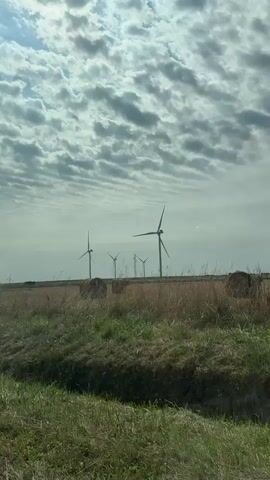 The video captures a landscape with several wind turbines in the distance, set against a cloudy sky. In the foreground, there is a grassy ditch and a field with hay bales scattered throughout. The wind turbines appear to be operational, with their blades slowly rotating. The overall scene suggests an agricultural or rural environment with a focus on renewable energy generation.