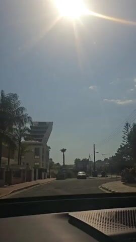 The video captures a view from inside a vehicle driving on a street under a bright, sunny sky. The sun is high and casts strong rays, creating lens flare. On the left side of the street, there's a modern building with an unusual, angled architectural design, accompanied by palm trees and other greenery. Further down the street, a single palm tree stands in the middle of a median. Several cars are visible on the road, with one car in the foreground and another in the distance. Utility poles with power lines run along the right side of the street, alongside trees and bushes. A stop sign is visible on the right, indicating a potential intersection or turn. The road appears to be paved, with clear lane markings. The overall environment suggests a warm, possibly Mediterranean, climate. The perspective is from the driver's or passenger's seat of a car, with the dashboard partially visible in the foreground.